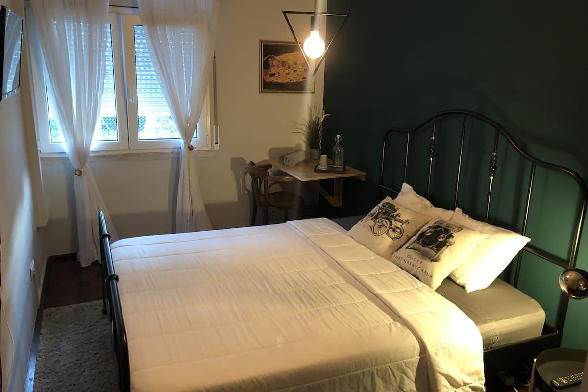 Een opgemaakt bed met witte lakens, een bureau met stoel, een wandlamp en een ingelijste foto.