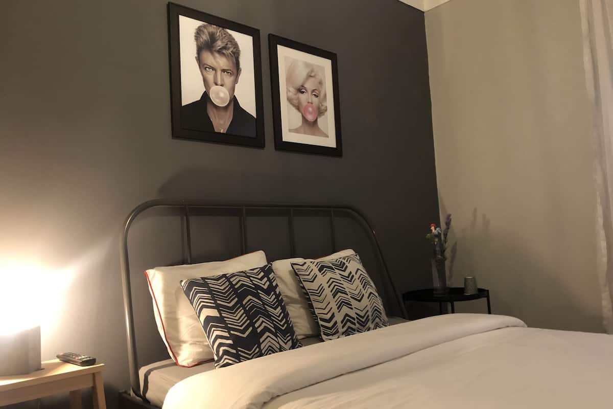 Een slaapkamer met een bed, kussens, een nachtkastje en twee ingelijste foto’s aan de muur.