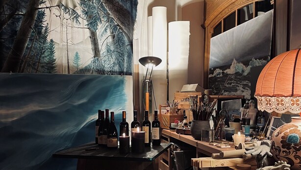 A cozy room with a large painting of a forest and waves, a table with wine bottles and a lamp, and a wooden desk with various items. - Hotel Sayonara (Folgaria)