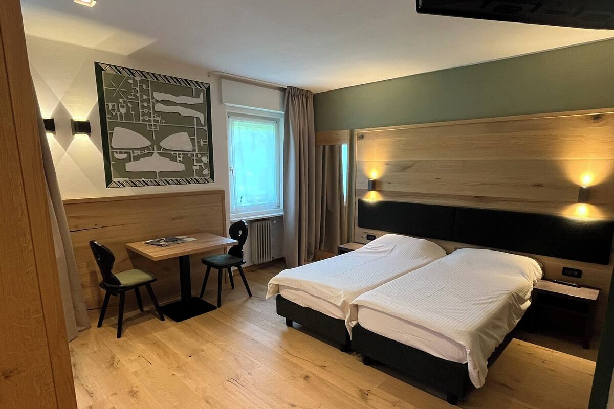 A hotel room with a wooden floor, a desk with a chair, a bed with two pillows, and a wall-mounted light fixture.