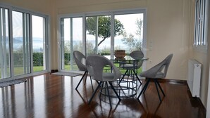 A dining area with a table and four chairs, large windows with a view of greenery and hills, and a radiator on the wall. - Villa Konstantina (Nafplio)