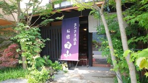 A traditional Japanese building with a stone entrance, a purple banner with white text, and lush greenery. - Kinosaki Onsen Ryokan Oyado Hakusan (Toyooka)