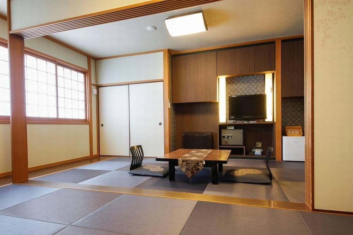 Ein traditioneller japanischer Raum mit Tatami-Bodenbelag, einem niedrigen Tisch mit Kissen, einem Fernseher und einer Küchenzeile.