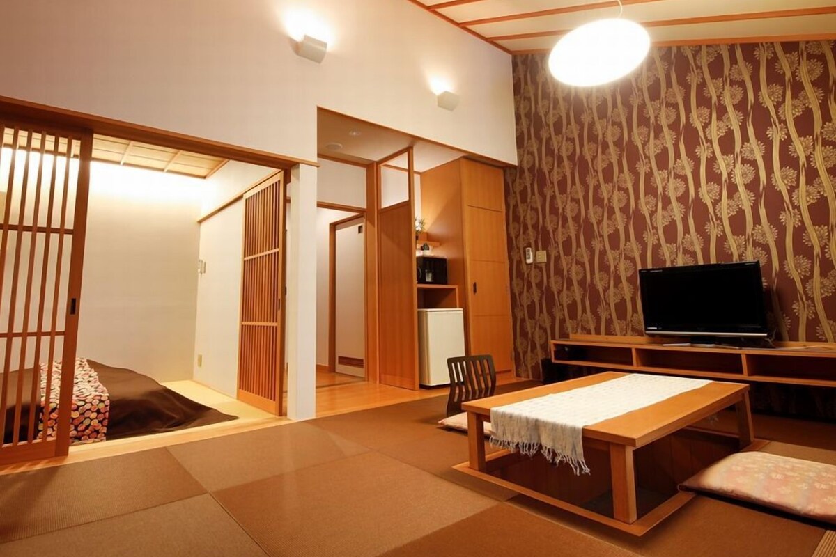 A traditional Japanese room with tatami flooring, a low table with a white cloth, a television, and sliding doors.