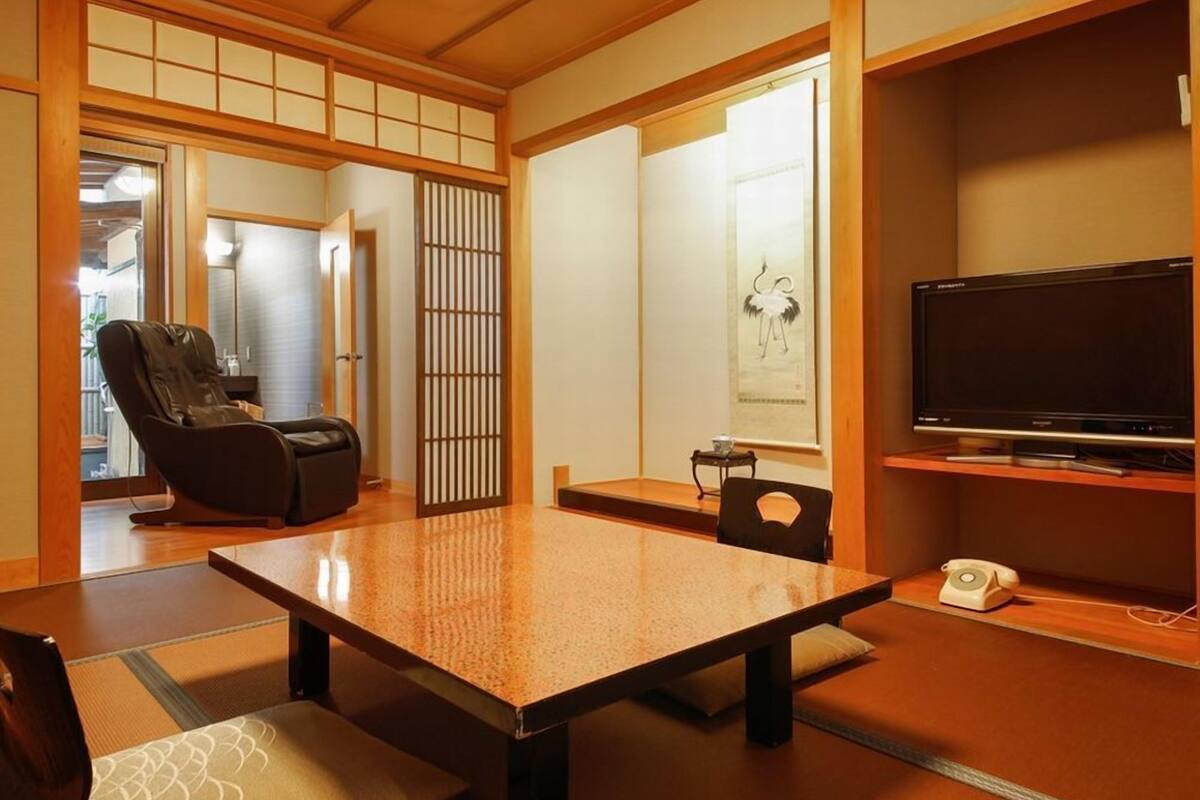 Ein traditioneller japanischer Raum mit Tatami-Boden, einem Tatami-Tisch, einem Massagesessel, einem Fernseher und einem Shoji-Bildschirm.