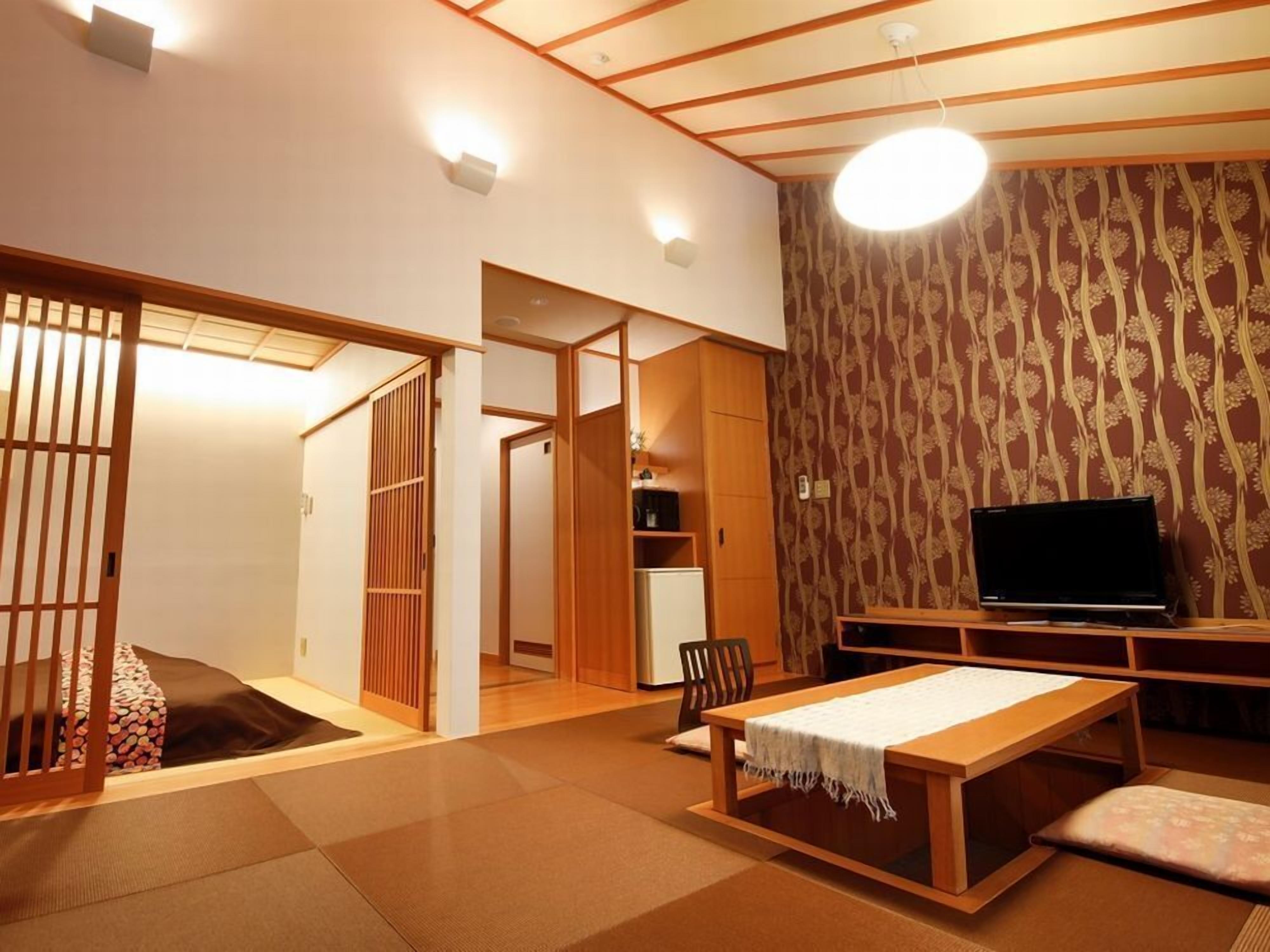 Une pièce de style japonais traditionnel, avec un sol recouvert de tatamis, une petite table recouverte d’une nappe blanche, un téléviseur posé sur un meuble en bois et une porte coulissante donnant sur une autre pièce.