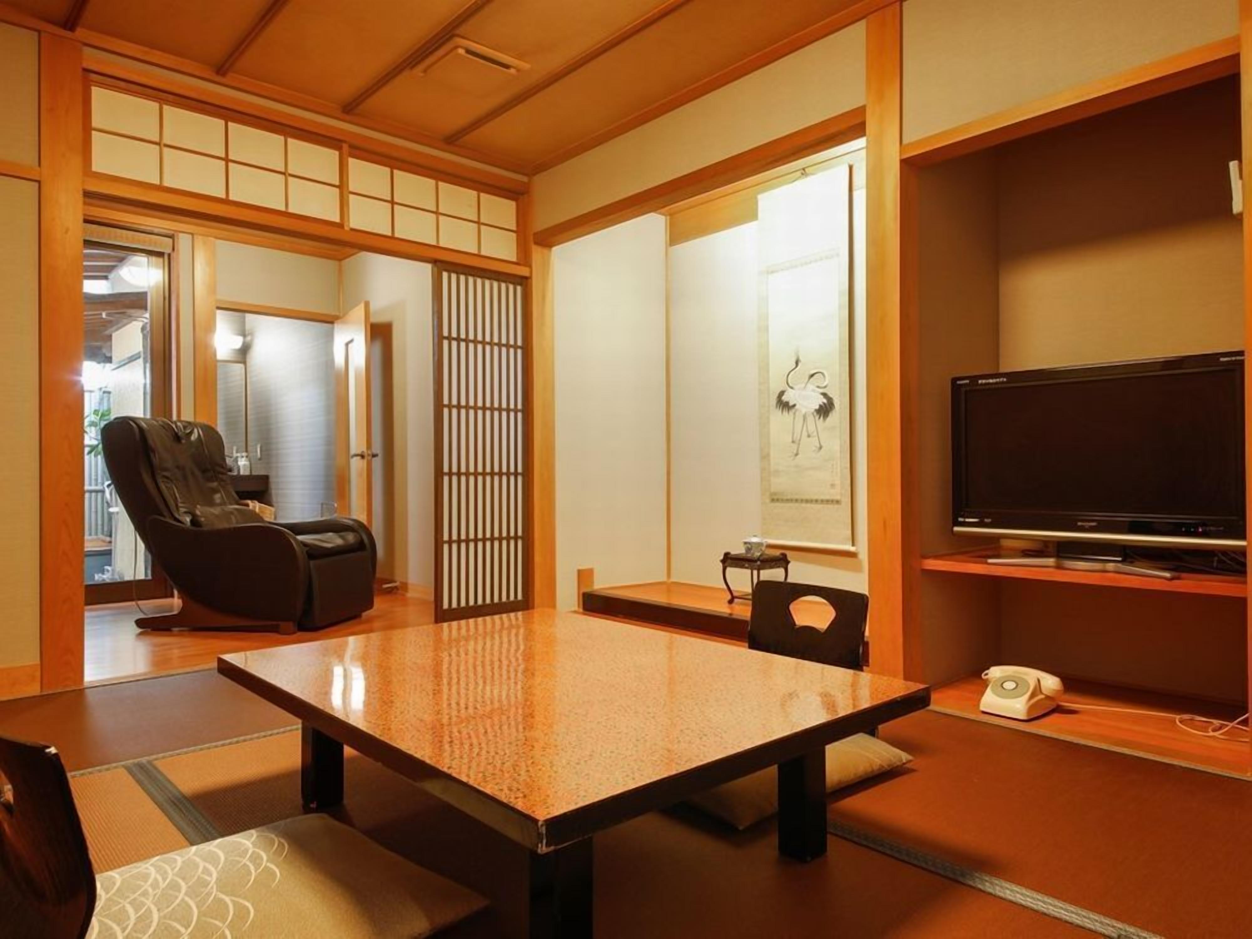 Une pièce de style japonais traditionnel, avec un sol recouvert de tatamis, une table en tatami, un fauteuil en cuir noir, un téléviseur posé sur un meuble en bois et un paravent shoji.