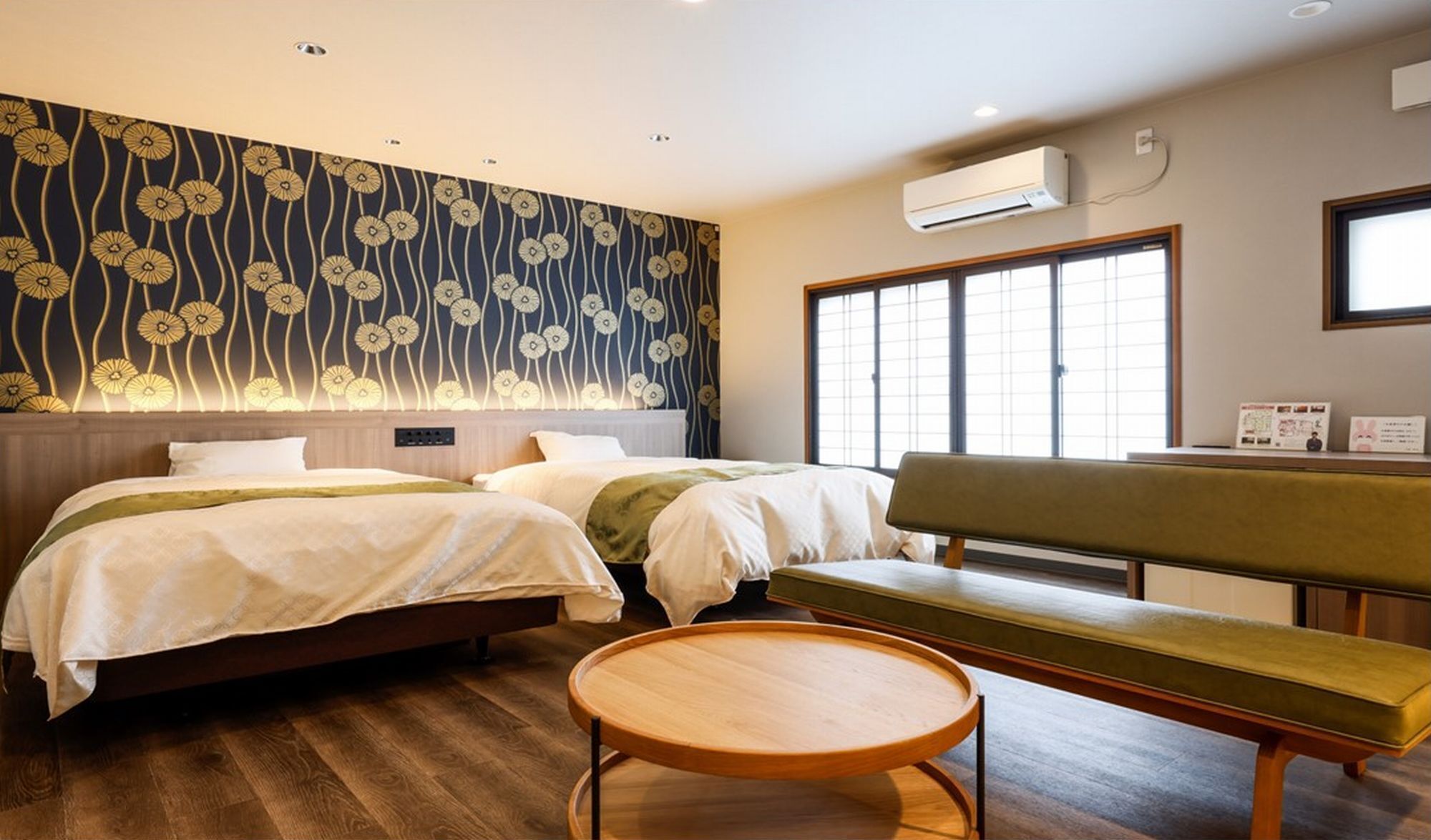 Habitación de hotel con dos camas, un banco, una mesa redonda de madera y un ventanal grande.