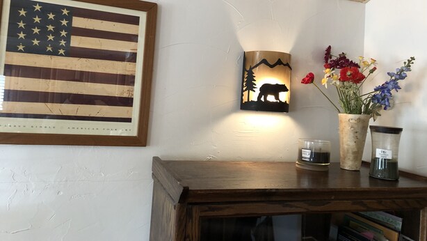 Un meuble en bois orné d’un drapeau américain encadré, un luminaire mural représentant la silhouette d’un ours, et un vase rempli de fleurs.