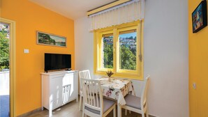 Une pièce avec un téléviseur posé sur un meuble blanc, une table à manger avec des chaises, une fenêtre donnant sur un paysage verdoyant et un tableau accroché au mur.
