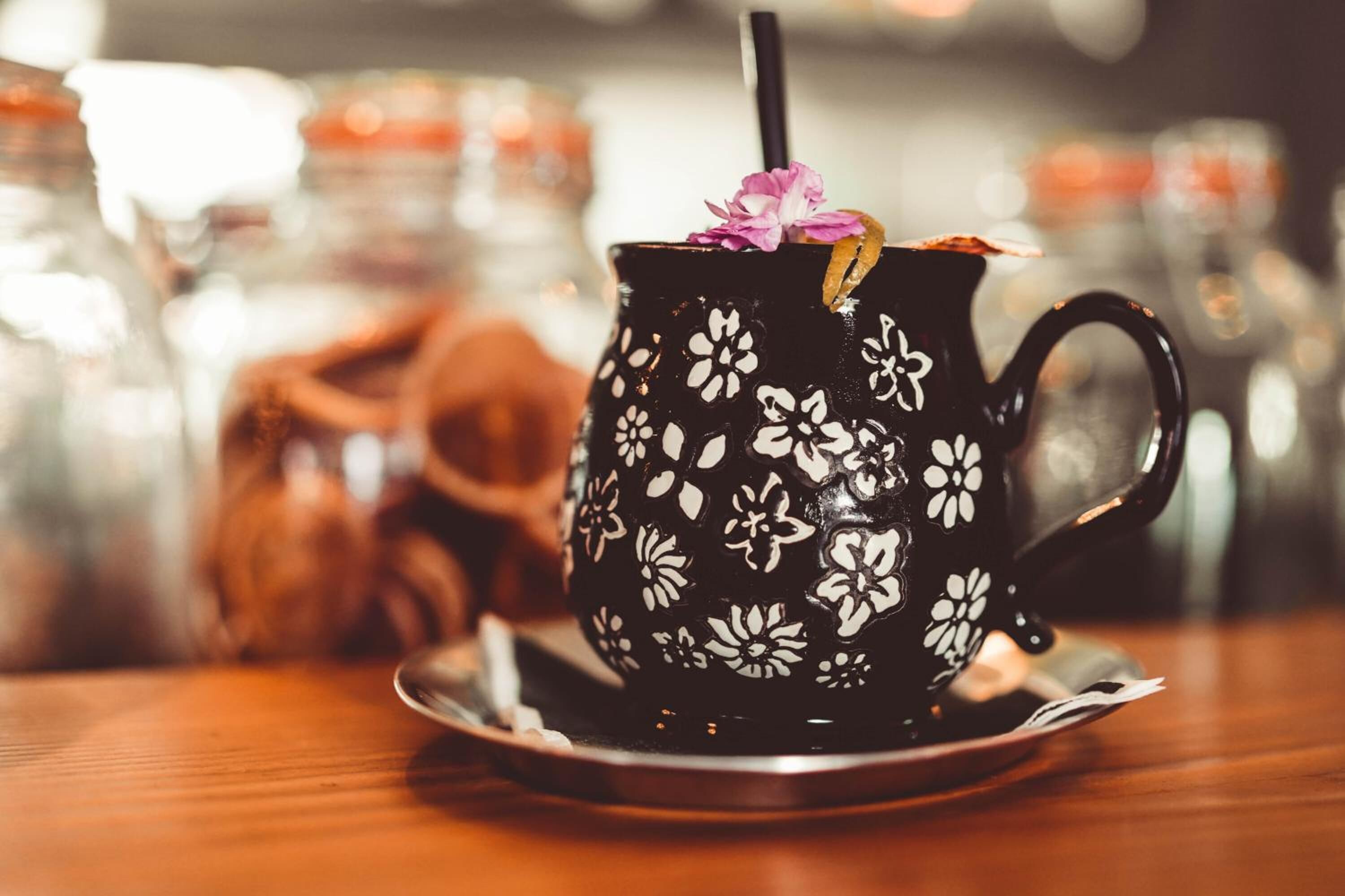 A black mug with white floral patterns, a straw, and a pink flower on top, placed on a saucer.