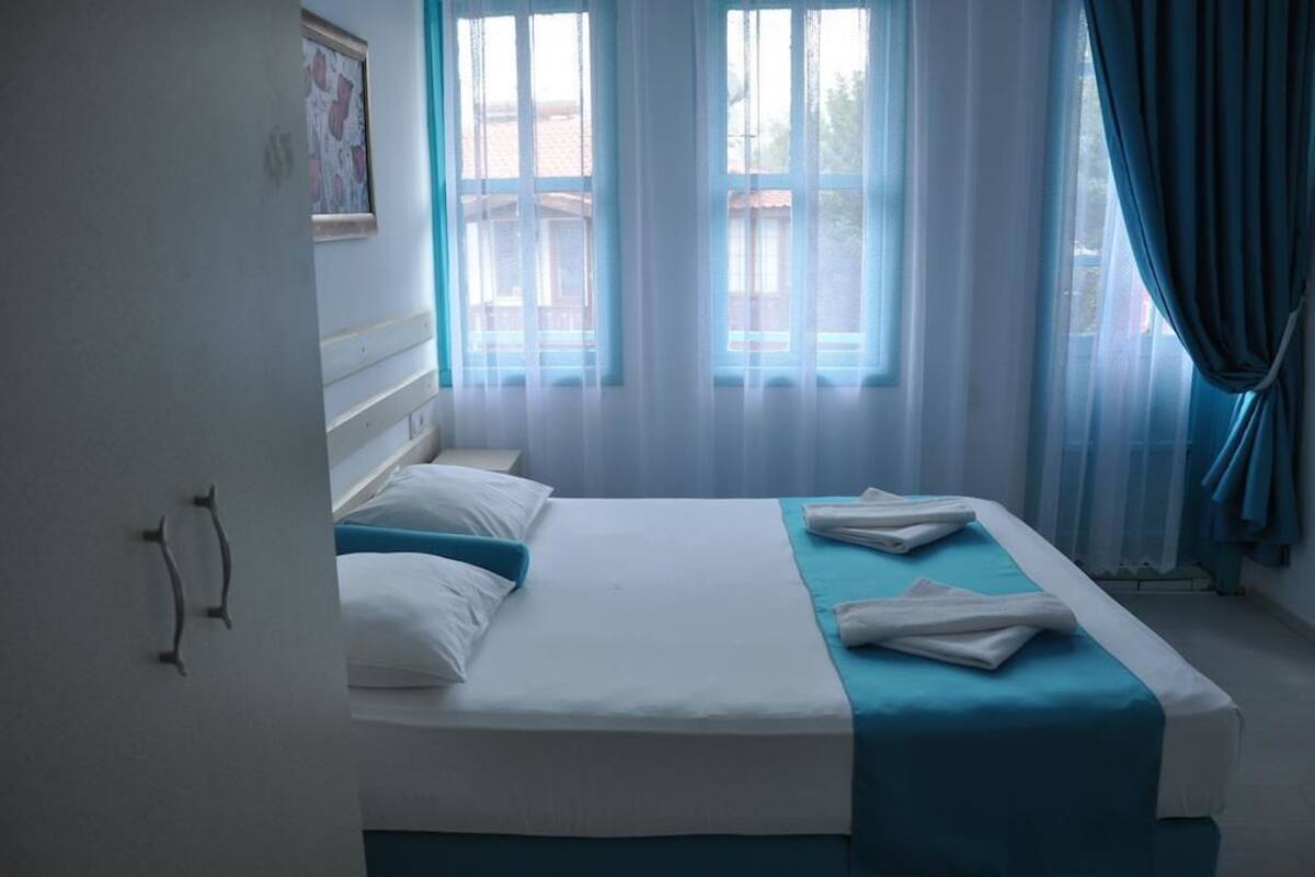 A neatly made bed with white and blue bedding, a window with sheer curtains, and a view of the outside.