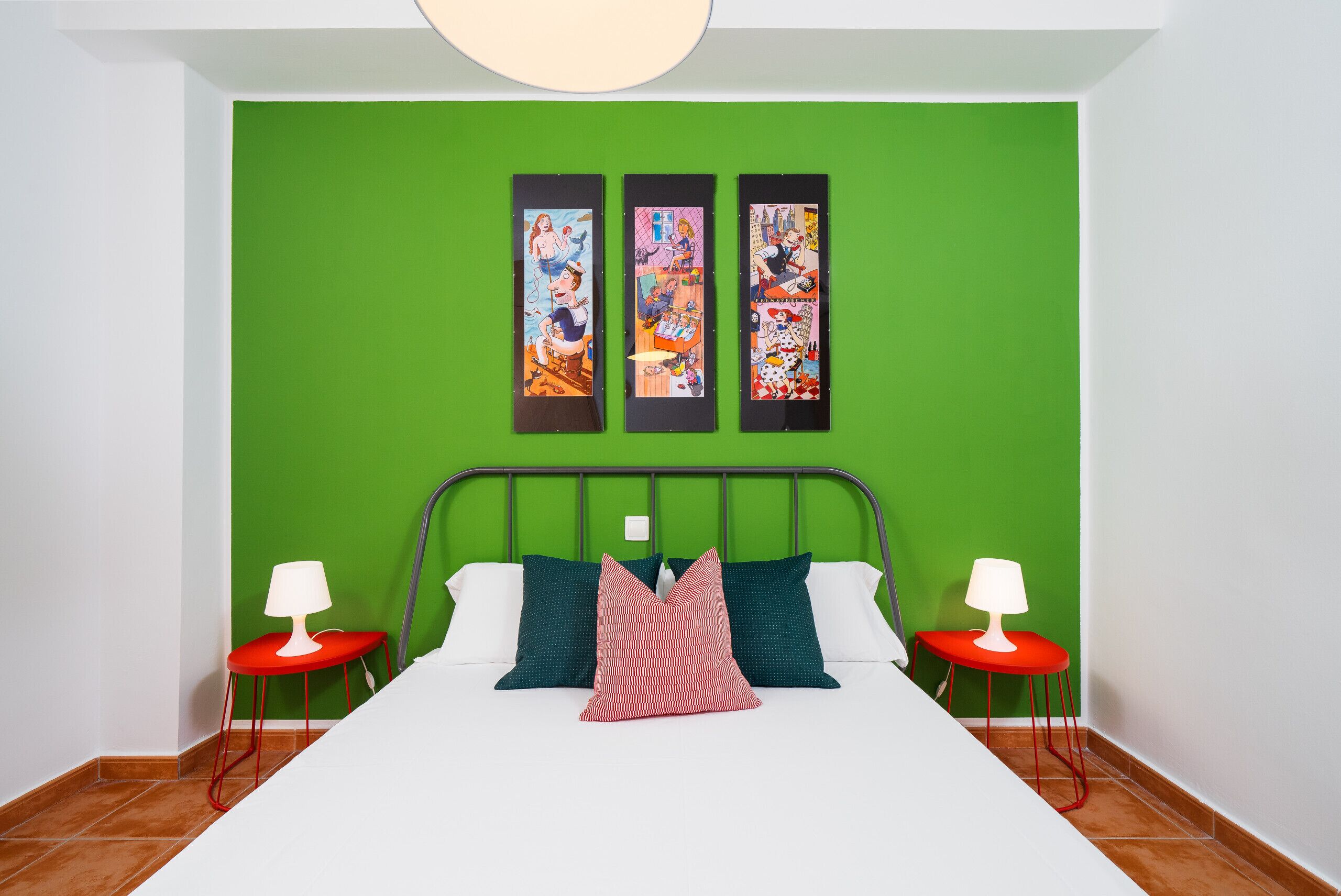 Een slaapkamer met een groene wand, een bed met wit beddengoed, twee rode nachtkastjes en drie ingelijste kunstwerken boven het bed.