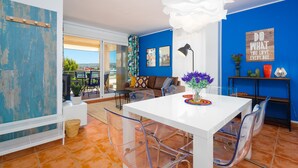 A modern living room with a white table, clear chairs, a sofa, and a blue accent wall. - Augusta Apartment Jávea Sur, Stylish with AC, Wifi, Terrace and Pool (Javea / Xàbia)