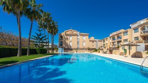 A swimming pool with a clear blue water, surrounded by palm trees and a building with balconies. - Augusta Apartment Jávea Sur, Stylish with AC, Wifi, Terrace and Pool (Javea / Xàbia)