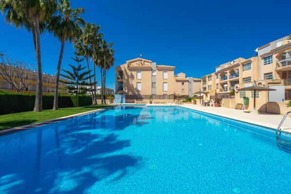 A swimming pool with a clear blue water, surrounded by palm trees and a building with balconies.