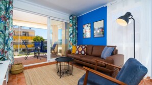 A living room with a brown sofa, a blue armchair, a small round table, and a floor lamp. There is a large window with a view of a building outside. - Augusta Apartment Jávea Sur, Stylish with AC, Wifi, Terrace and Pool (Javea / Xàbia)