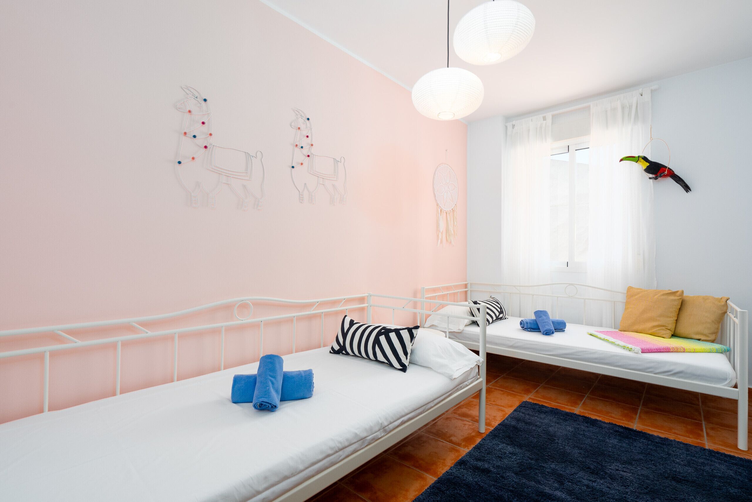 A room with two beds, a window with sheer curtains, a hanging birdcage, and wall art.