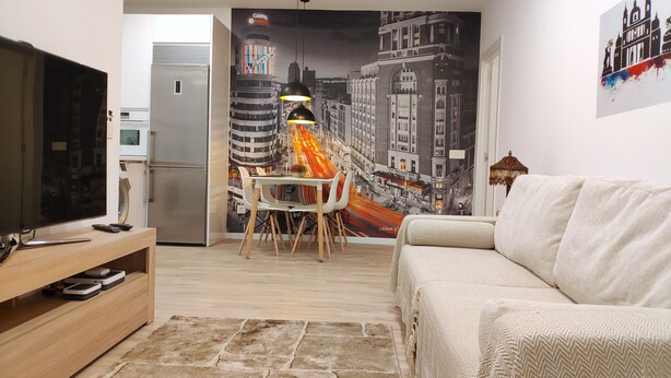 Un salon équipé d’un téléviseur à écran plat, d’un canapé, d’un coin repas avec une table et des chaises, d’un réfrigérateur et d’une fresque murale représentant un paysage urbain.