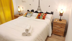 Un lit bien fait, agrémenté de serviettes pliées, un mur décoré d’une fresque représentant un paysage urbain et deux lampes de chevet.