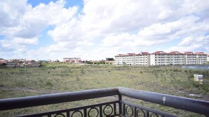 A view from a balcony with a railing, overlooking a grassy area and a large building with multiple floors and red roofs. - Joy Comfort Homestay (Syokimau)