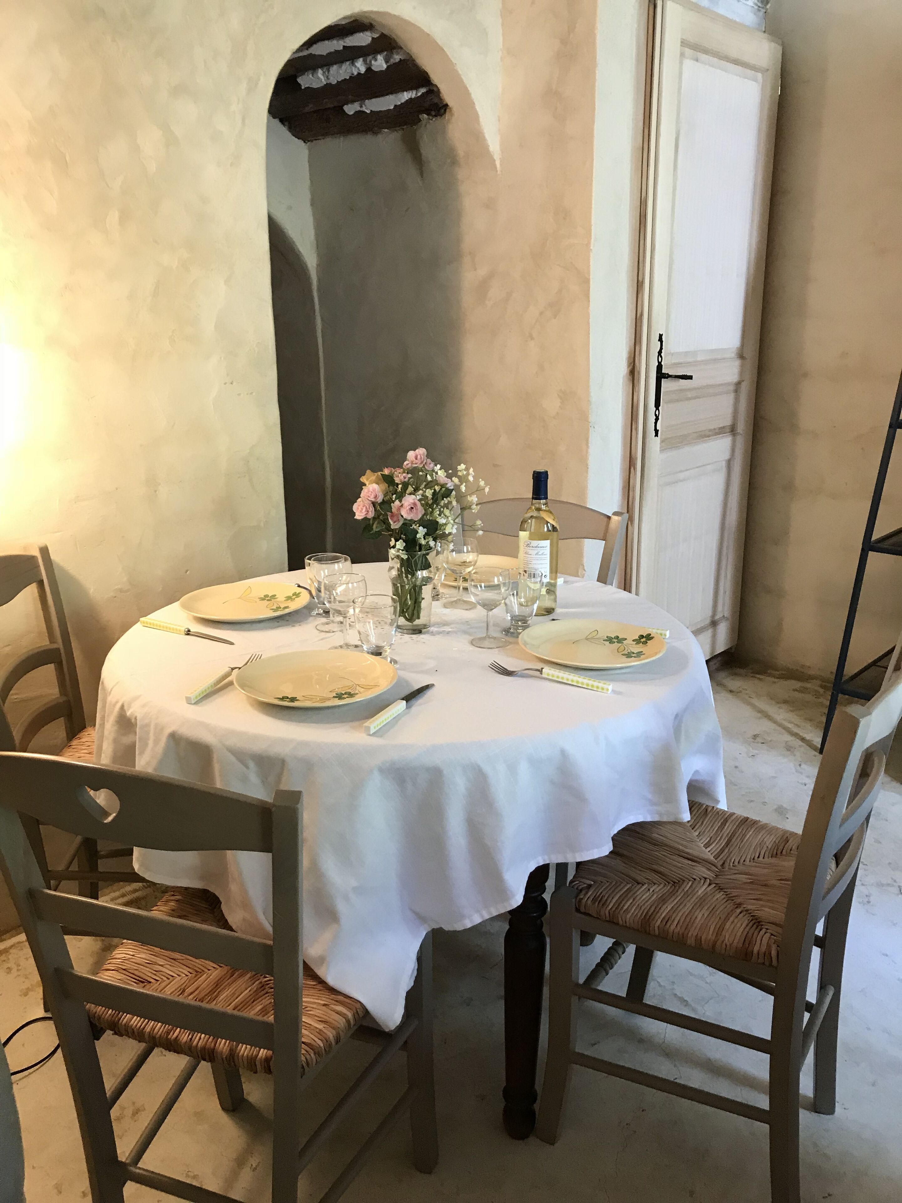 A round dining table with a white tablecloth, surrounded by four wooden chairs with woven seats. A vase with flowers and a bottle of wine are placed on the table.