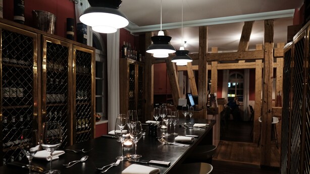 Una larga mesa de comedor dispuesta con copas de vino, cubiertos y velas, con interior de madera y lámparas colgantes.