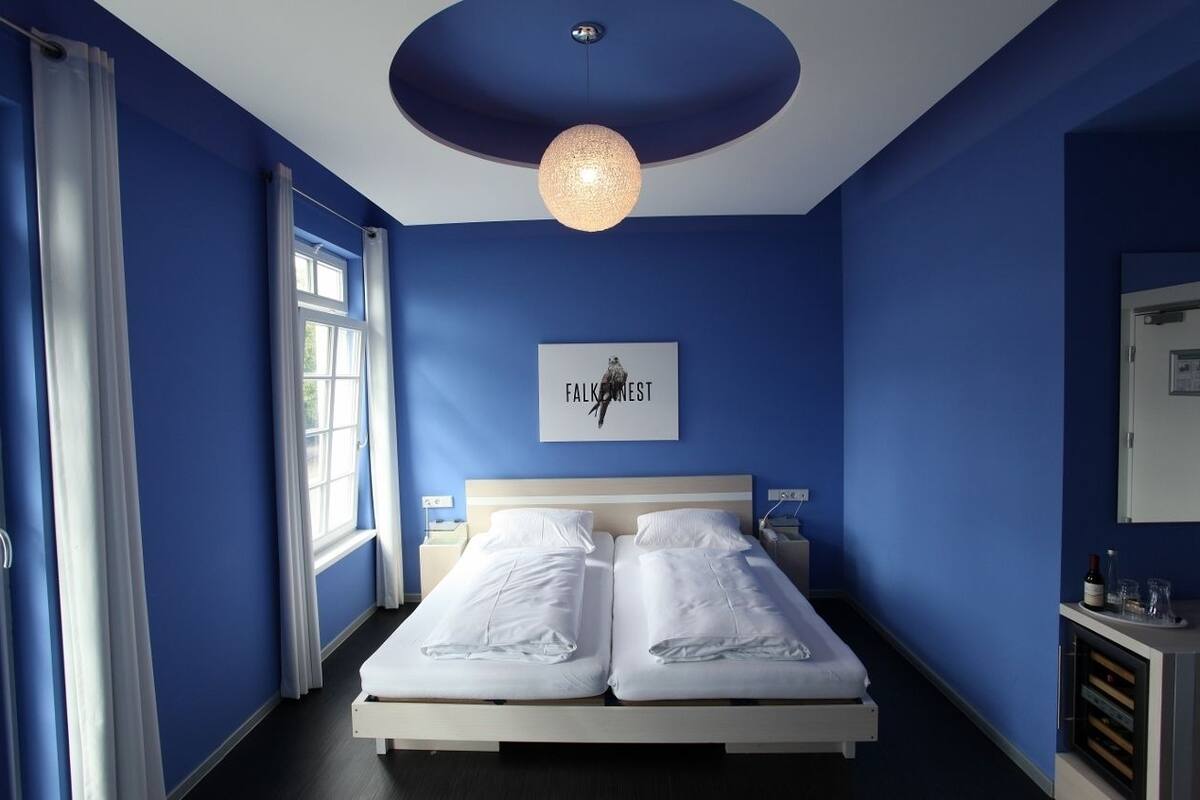 A bedroom with two beds, a large window with curtains, a ceiling light, and a wall-mounted artwork.