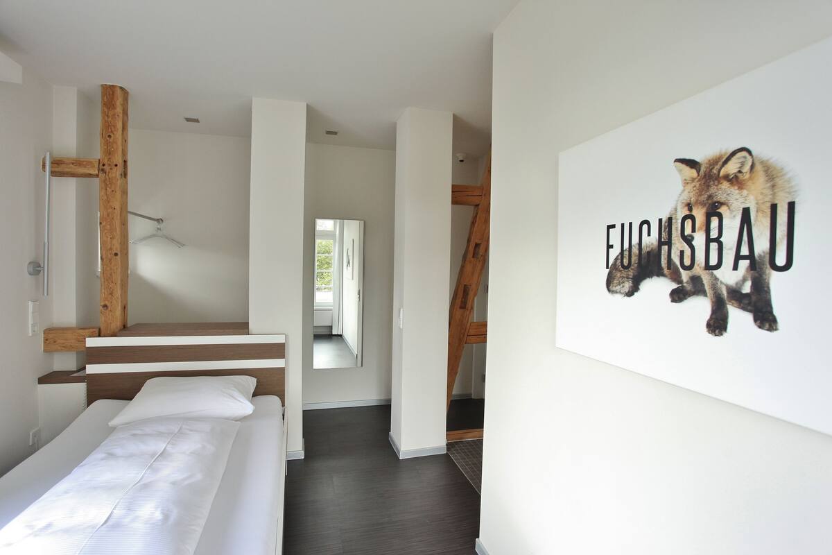 A modern hotel room with a wooden bed, a white wall with a fox illustration, and a view of the outdoors.