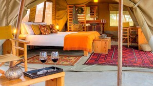 Un hébergement confortable en forme de tente, comprenant un lit, une petite table en bois avec des verres de vin rouge, et une autre petite table sur laquelle est posé un panier en osier.