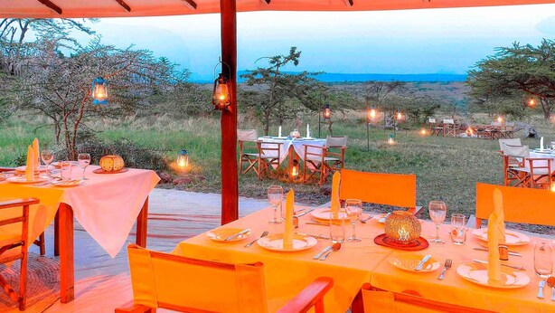 Un espace de restauration en plein air, avec des tables disposées pour deux personnes, entouré d’un cadre naturel et illuminé pour la soirée.