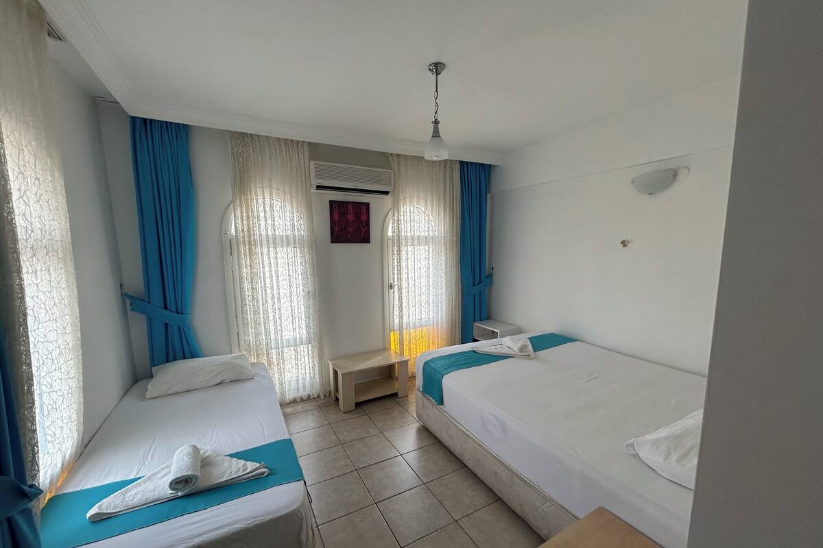 Une chambre d’hôtel avec deux lits simples, une petite table, un ventilateur de plafond et une fenêtre avec des rideaux.