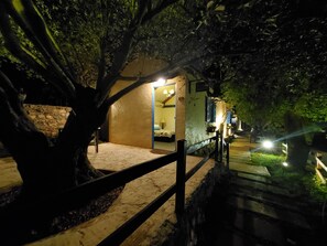 Uma área externa rústica, com cerca de madeira, parede de pedra e um caminho que leva a uma entrada iluminada de um edifício.