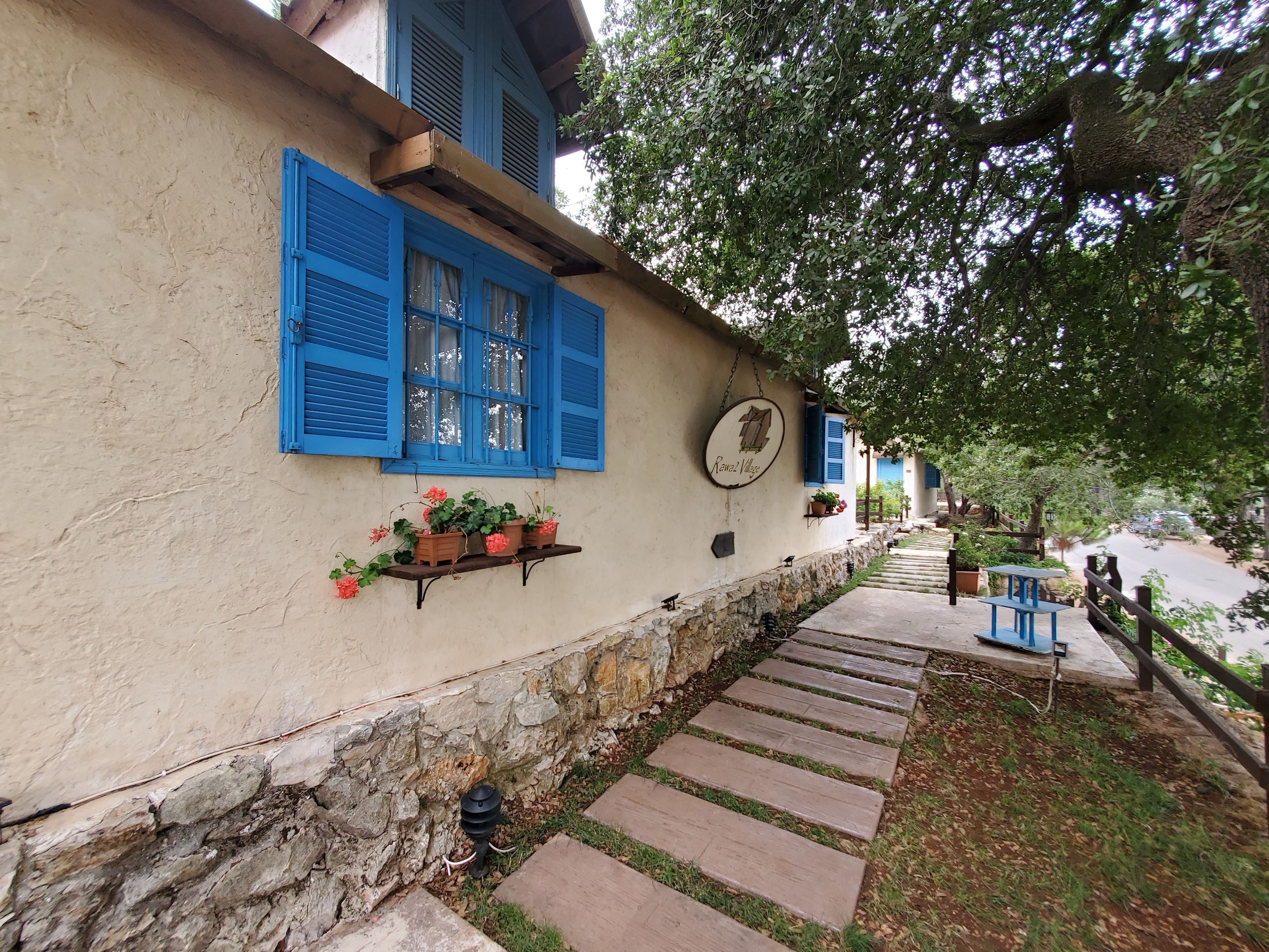 A rustic building with blue shutters, a stone wall, and a wooden pathway leading to a bench.