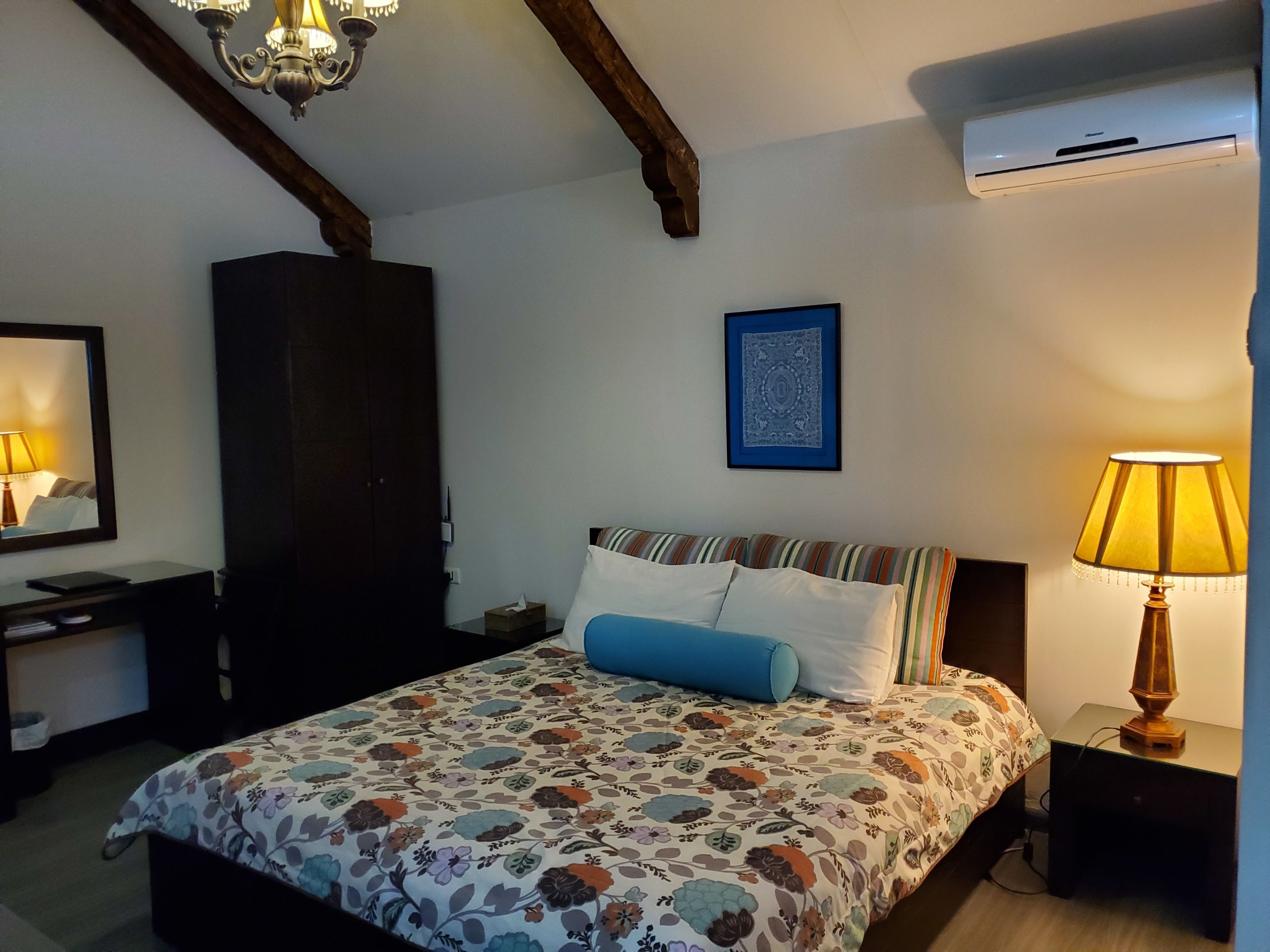 A bedroom with a bed, a nightstand, a dresser, a mirror, a chandelier, and an air conditioning unit.