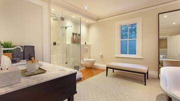 A modern bathroom with a glass shower, a vanity with a marble countertop, a bench, and a window with a view of trees.