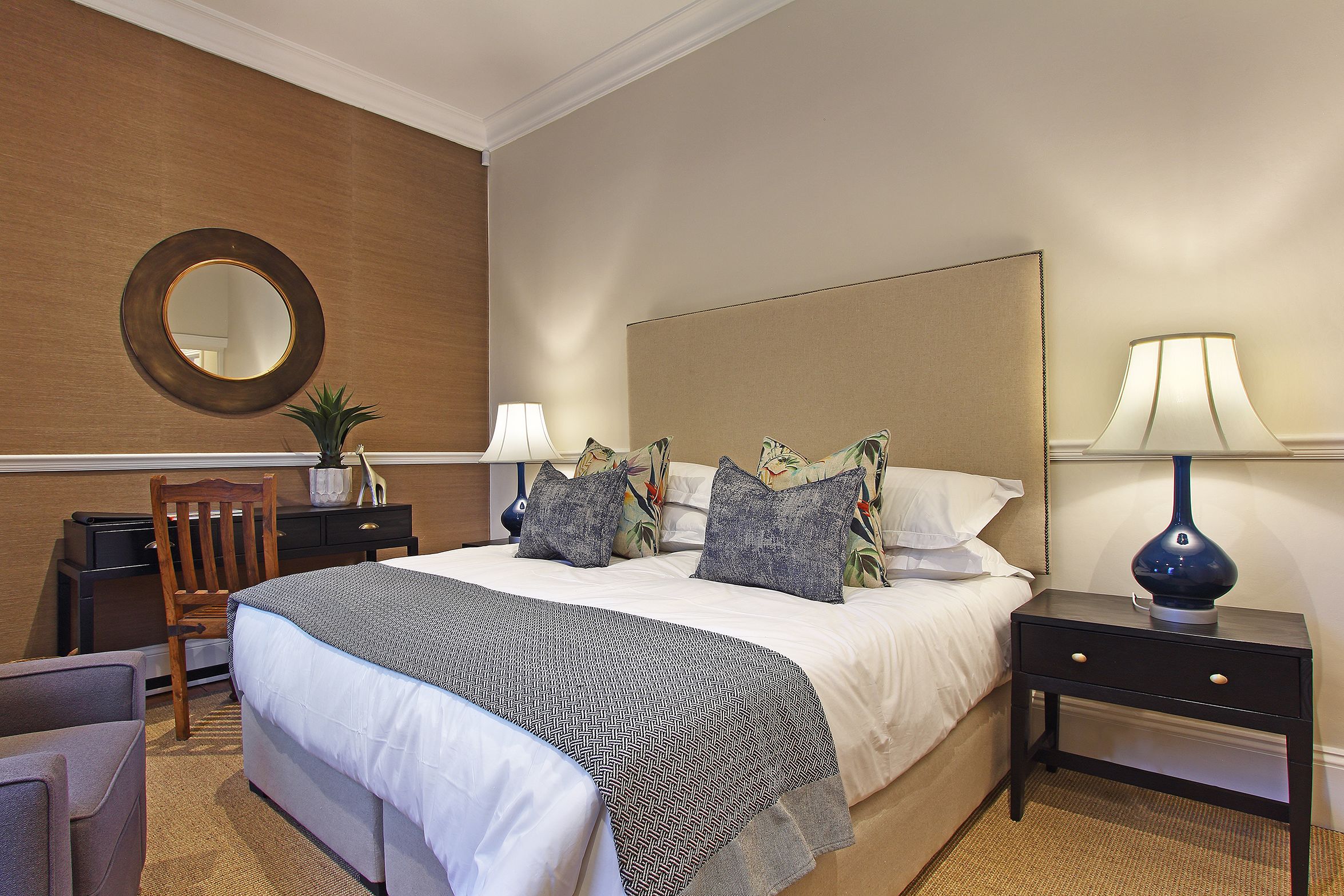 Quarto de hotel bem arrumado com uma cama grande, mesa de cabeceira com abajur, uma cadeira e uma pequena escrivaninha.