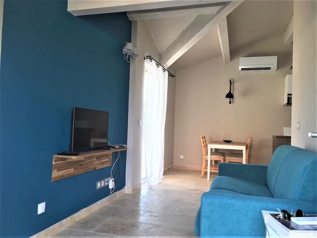 A living room with a blue sofa, a TV on a wooden shelf, a dining table with chairs, and a kitchen area with an air conditioner.