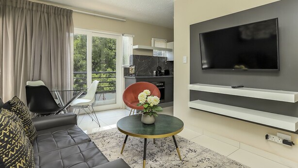 Un soggiorno moderno con un divano, un tavolo da pranzo con sedie, una TV a parete e una cucina visibile attraverso una porta aperta.