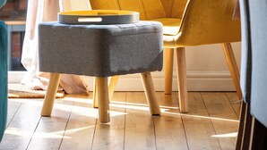 Una habitación con un sillón amarillo, un reposapiés gris y una ventana con cortinas.