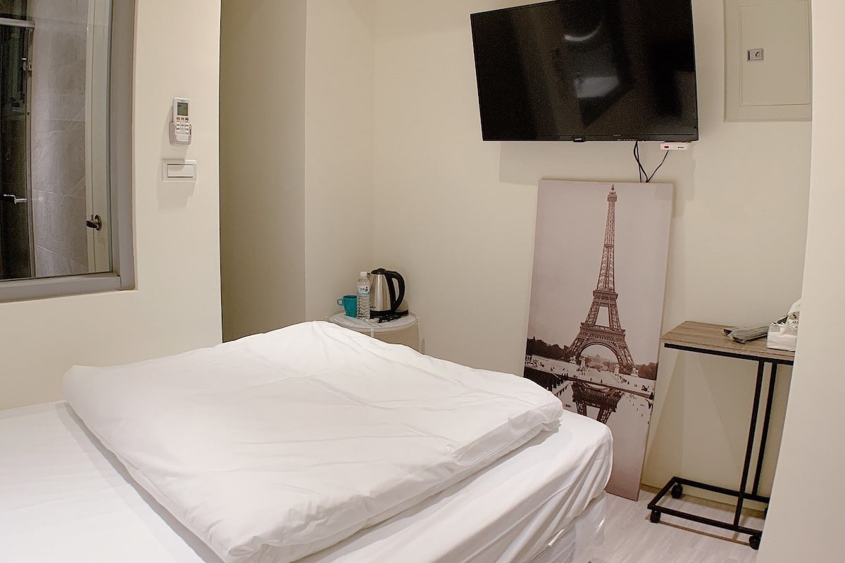 A hotel room with a bed, a flat-screen TV mounted on the wall, an air conditioning unit, a small table with a kettle, a cup, and a bottle, and a poster of the Eiffel Tower.