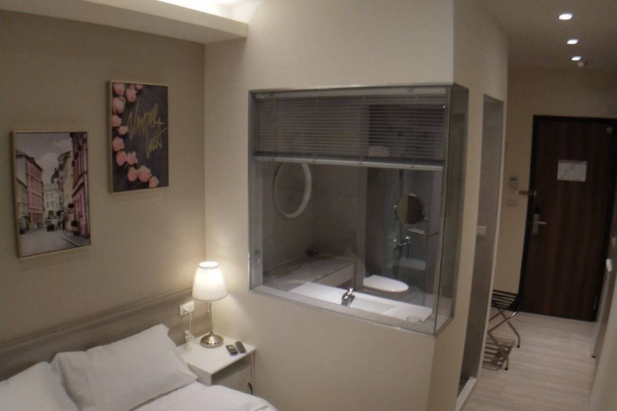 A hotel room with a bed, a nightstand with a lamp, a bathroom with a window, and a door.