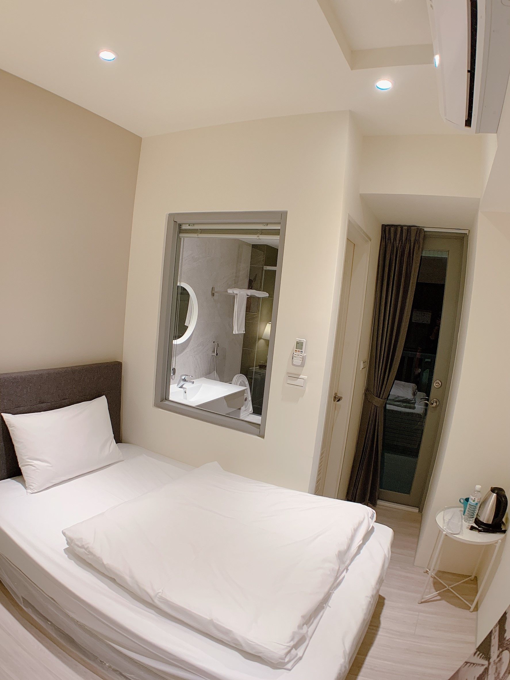 A hotel room with a bed, a bedside table with a lamp, a bathroom visible through a glass door, and a ceiling light.