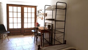 Een kamer met een houten deur, een metalen stapelbed, een tafel met stoelen en een klein plankje.