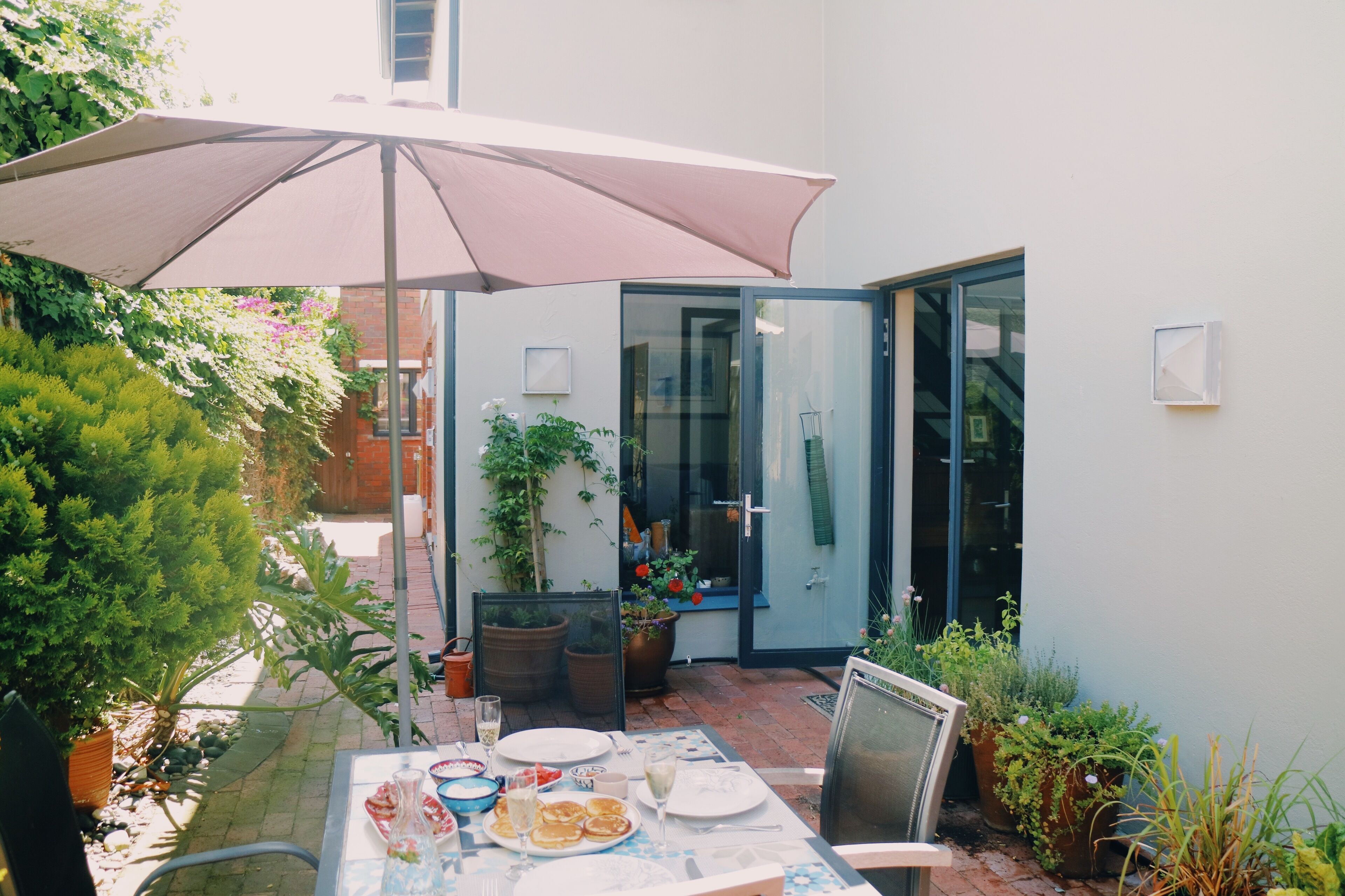 En udendørs patio med et spisebord, omgivet af krukkeplanter og en stor parasol.