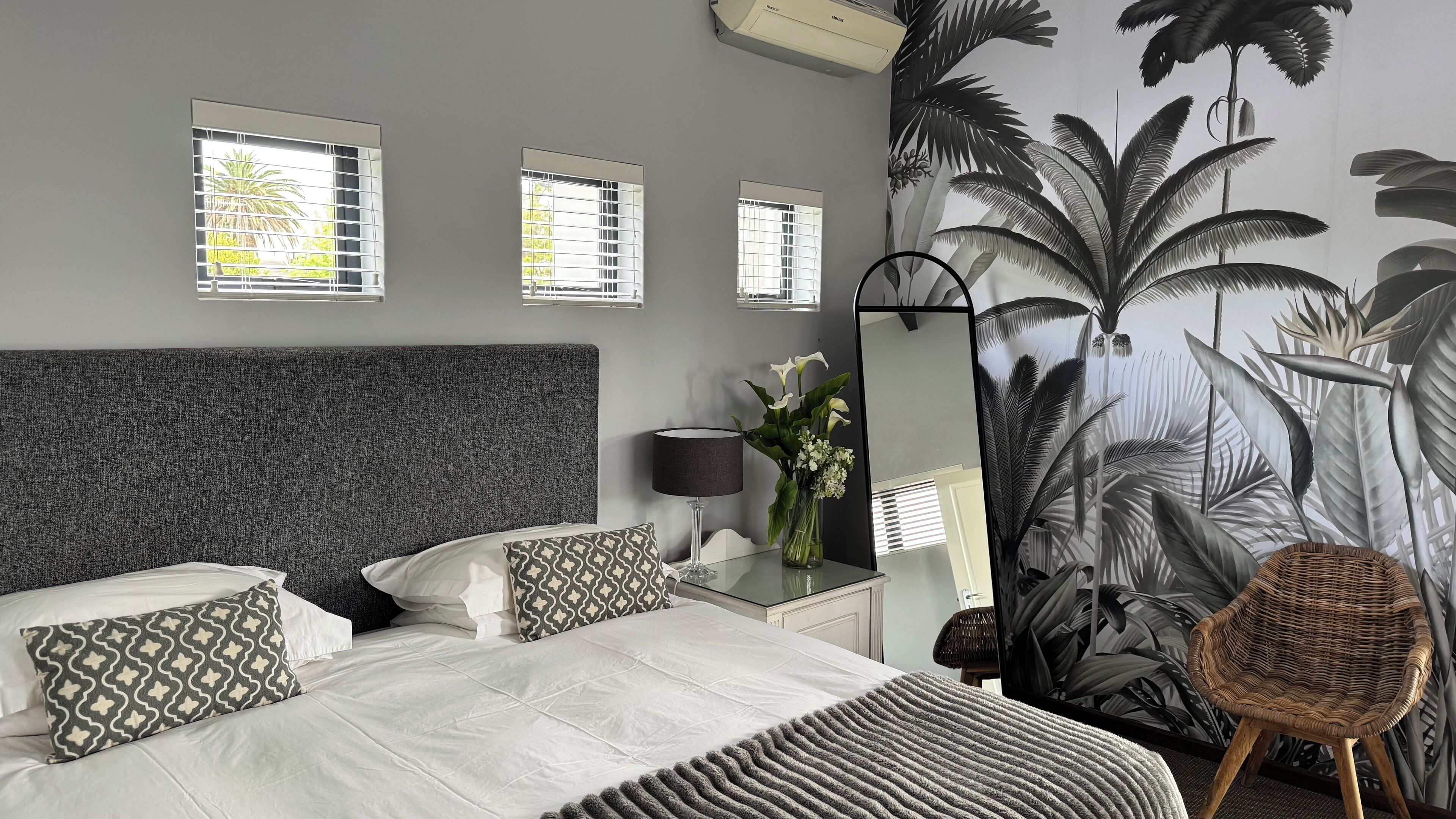 A bedroom with a grey headboard, a bed with white bedding, patterned pillows, a bedside table with a lamp, a mirror, and a wall with a tropical plant mural.