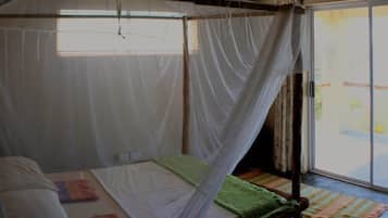 A room with a bed under a mosquito net, a small table, and a view of the outside through a sliding door.