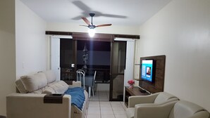 Ein Wohnzimmer mit Sofa, Sessel, Fernseher, Deckenventilator und Blick auf den Balkon.