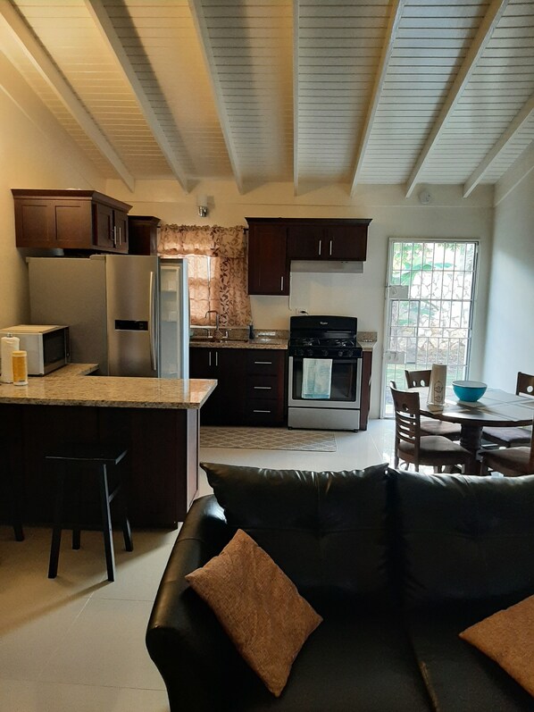 A modern kitchen with dark wooden cabinets, a stainless steel refrigerator, and a gas stove. There is a dining area with a table and chairs, and a black leather sofa with brown cushions.