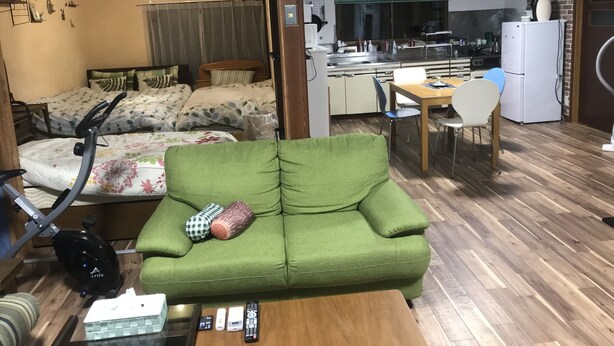 A compact living space with a green sofa, a bed with floral bedding, a dining table with chairs, a kitchen area, and a small exercise bike. - Airfield hill and patchwork hill valley house (旭川市)